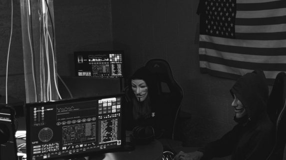 Two anonymous hackers in a dim room working on computers, symbolizing a cyber attack.